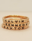 Three gold-colored stacking rings with the names CALVIN, KENNEDY, and PAXTON engraved in bold uppercase letters.