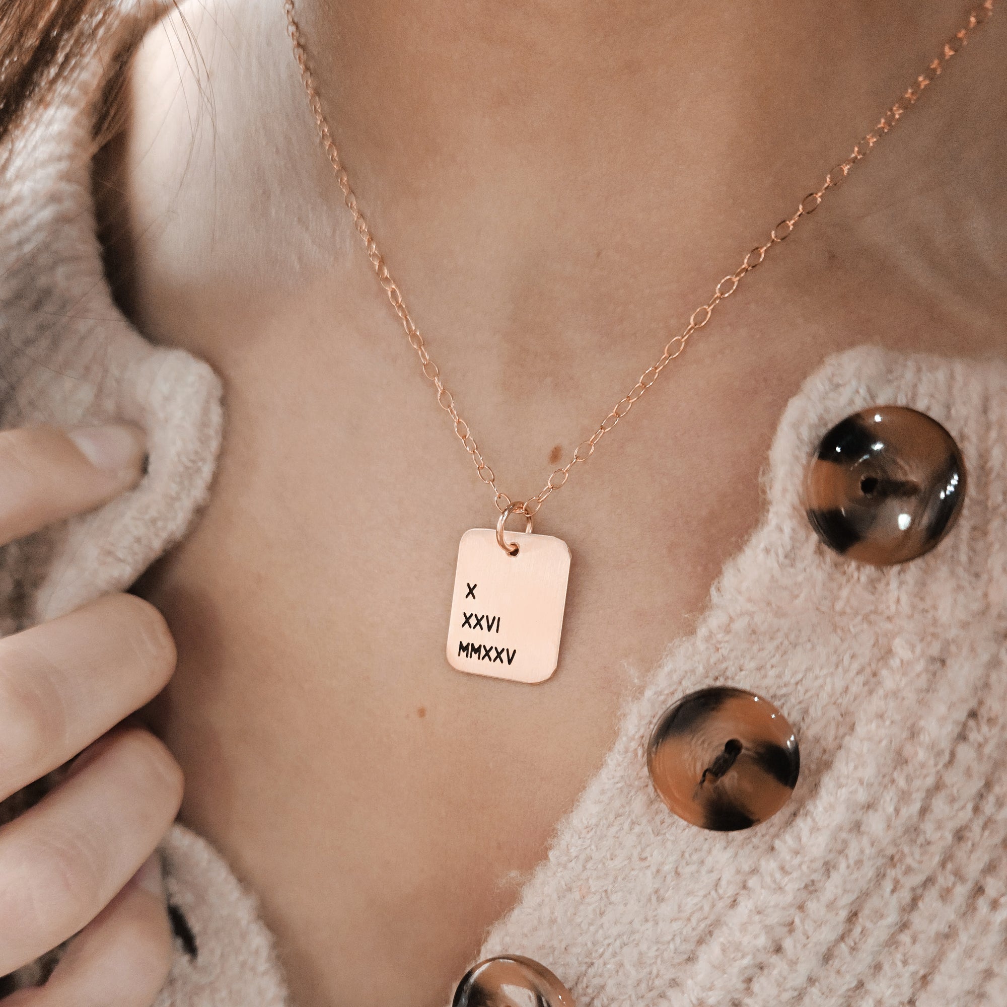 A person wearing a cardigan is showcasing a gold necklace with a rectangle pendant engraved with Roman numerals.