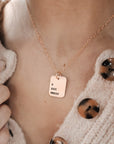 A person wearing a cardigan is showcasing a gold necklace with a rectangle pendant engraved with Roman numerals.