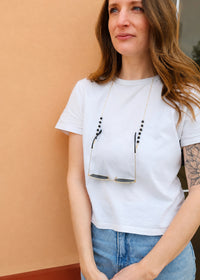 Woman wearing a white t-shirt with sunglasses hanging from her neckline against a beige wall.