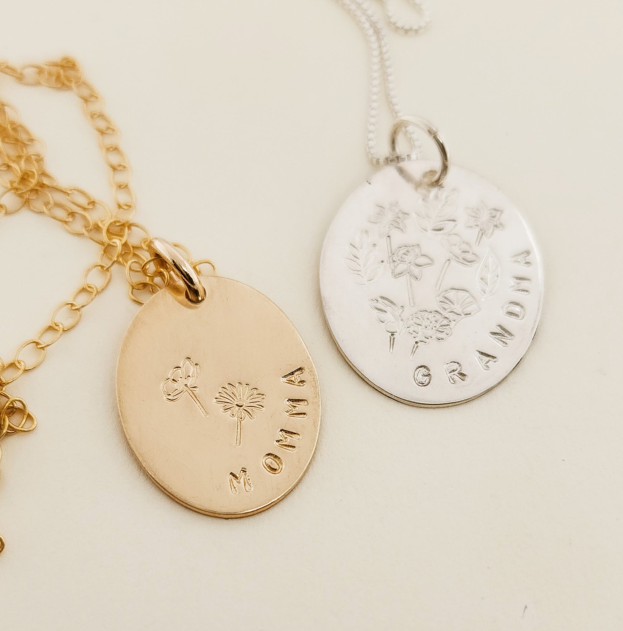 Two necklaces with gold-filled or silver round charms, one with a floral design and the other with a more detailed floral wreath design, attached to small gold or silver chains.