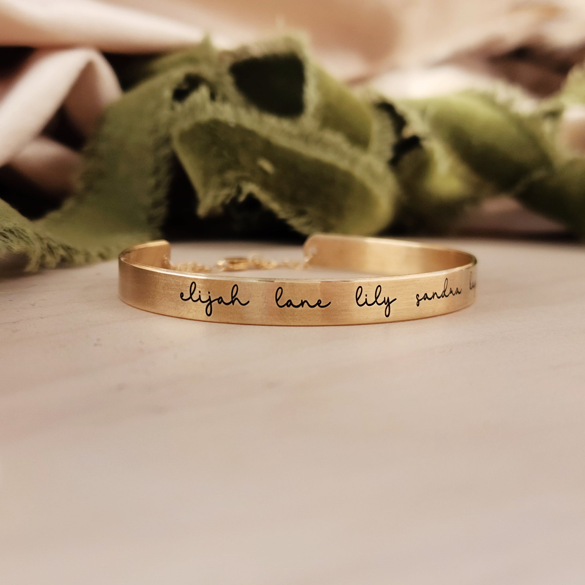 A gold-colored cuff bracelet with custom names engraved on it, displayed on a white surface with a green textured object to the side.