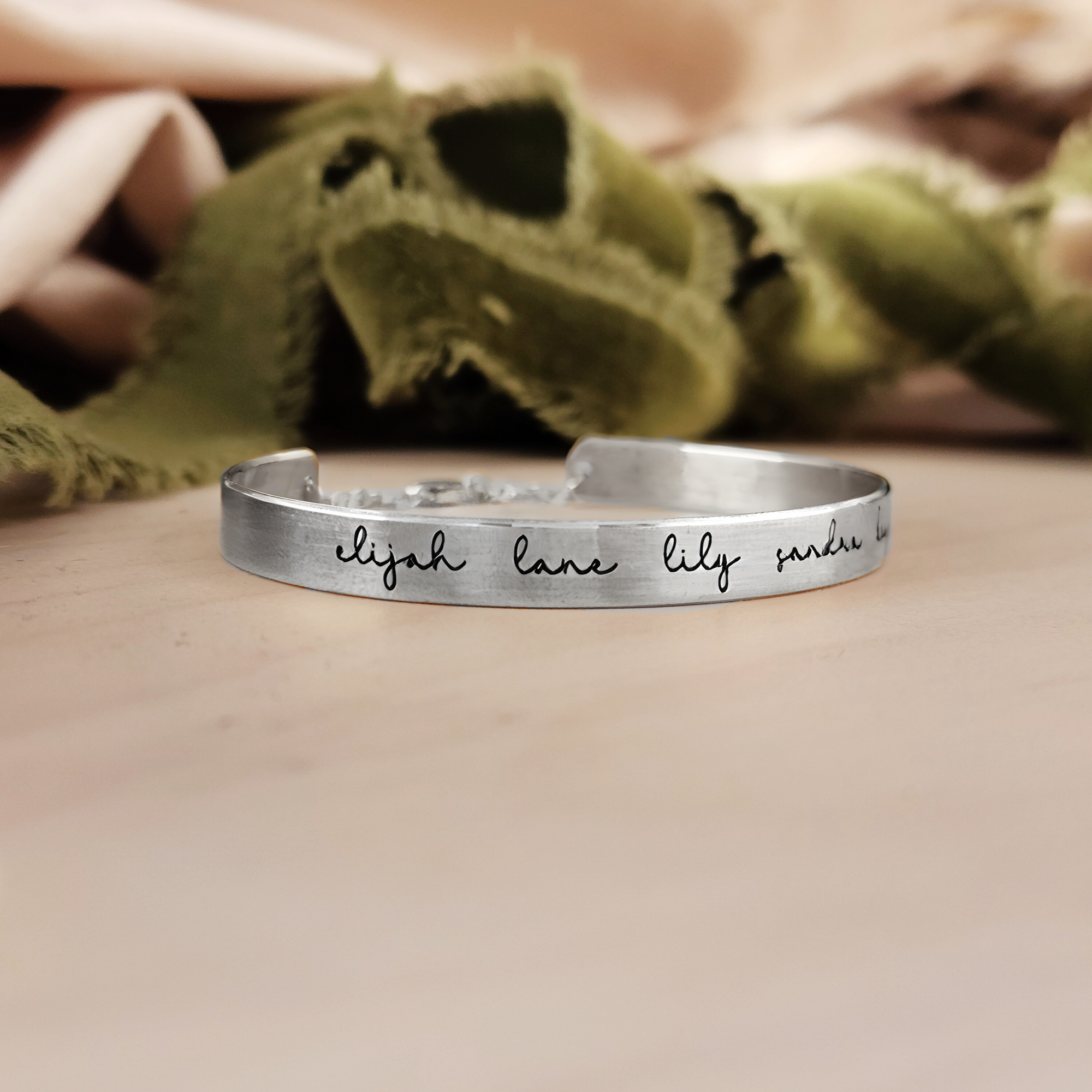 A gold-colored cuff bracelet with custom names engraved on it, displayed on a white surface with a green textured object to the side.