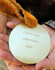 Child holding a 'Liam's First Christmas 2025' ornament with a teddy bear in the backgroung