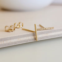 A pair of 14K yellow gold-filled sparkle bar earrings displayed on a wooden surface with a white circular object in the background.