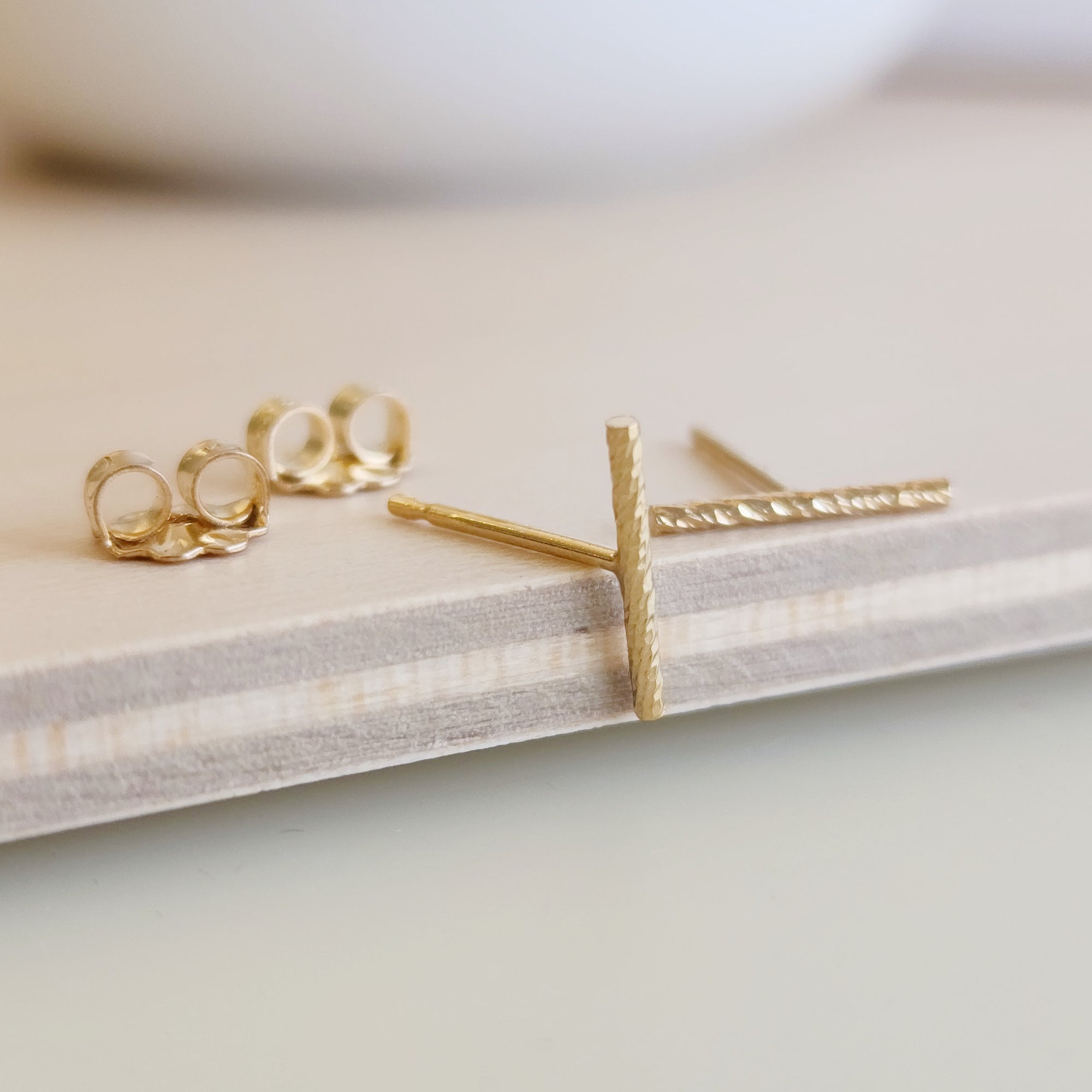 A pair of 14K yellow gold-filled sparkle bar earrings displayed on a wooden surface with a white circular object in the background.