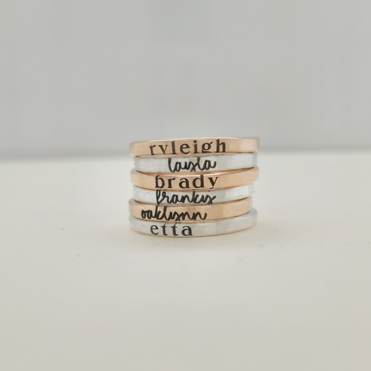There are six stacking name rings piled on a white table.  The bottom ring is a 2mm silver name ring with a serif font that says "etta."  Above it, is a 2mm yellow gold ring in cursive that says "oaklynn."  Then, above it, a ring that "franky"then "brady" then "layla" then "ryleigh."  The rings alternate from silver to gold.