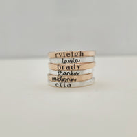 There are six stacking name rings piled on a white table.  The bottom ring is a 2mm silver name ring with a serif font that says "etta."  Above it, is a 2mm yellow gold ring in cursive that says "oaklynn."  Then, above it, a ring that "franky"then "brady" then "layla" then "ryleigh."  The rings alternate from silver to gold.
