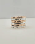 There are six stacking name rings piled on a white table.  The bottom ring is a 2mm silver name ring with a serif font that says "etta."  Above it, is a 2mm yellow gold ring in cursive that says "oaklynn."  Then, above it, a ring that "franky"then "brady" then "layla" then "ryleigh."  The rings alternate from silver to gold.