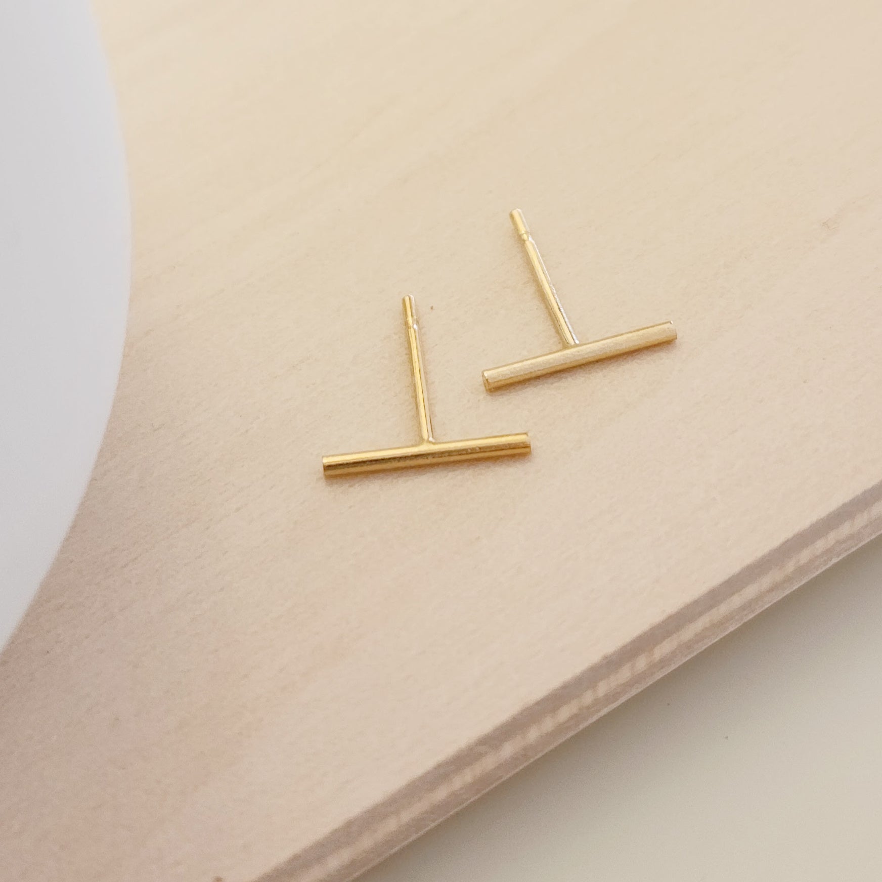 A pair of gold-colored bar earrings placed on a wooden surface next to a white cup.