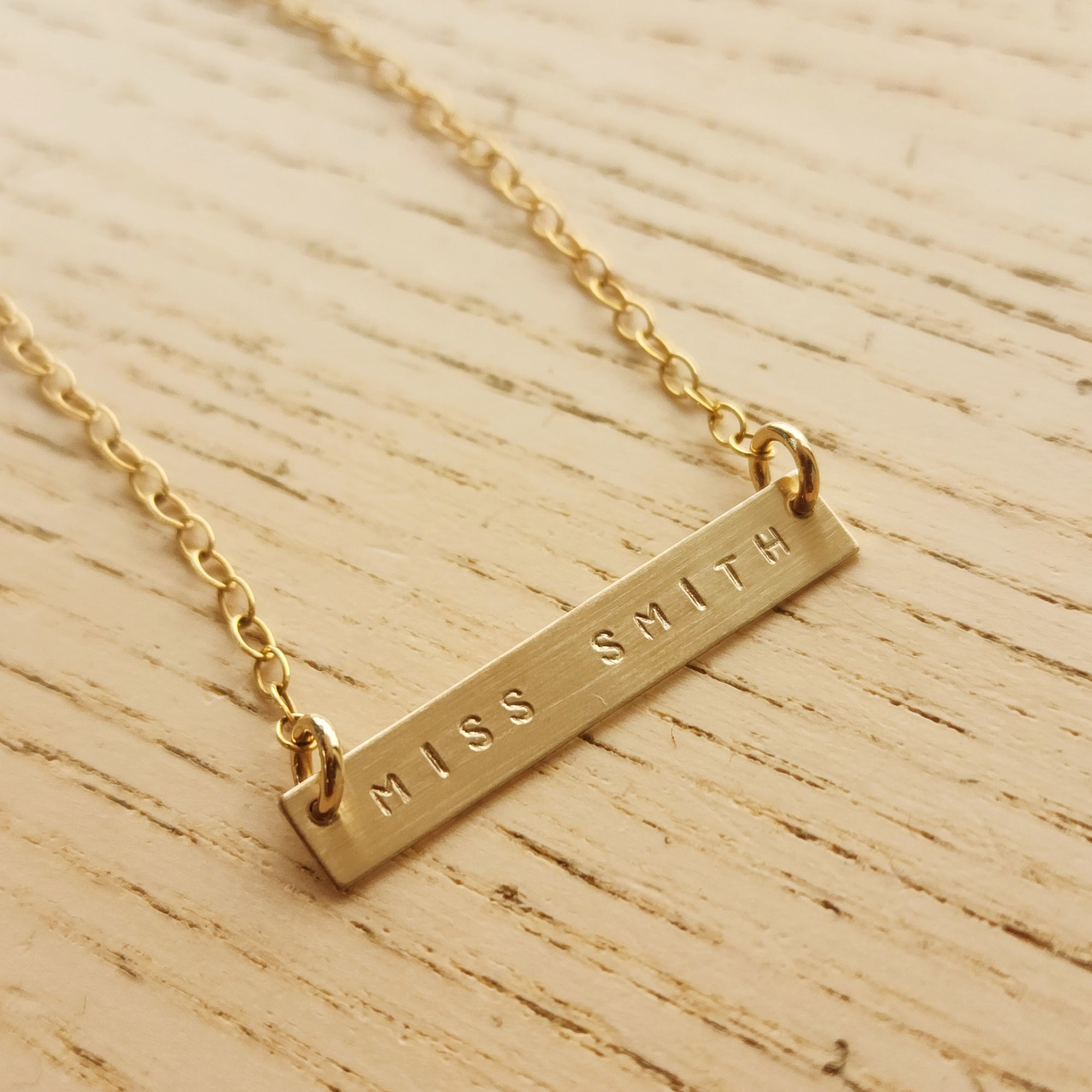 Close-up of brushed gold bar teacher necklace engraved with MISS SMITH, displayed on a light wood background with cable chain