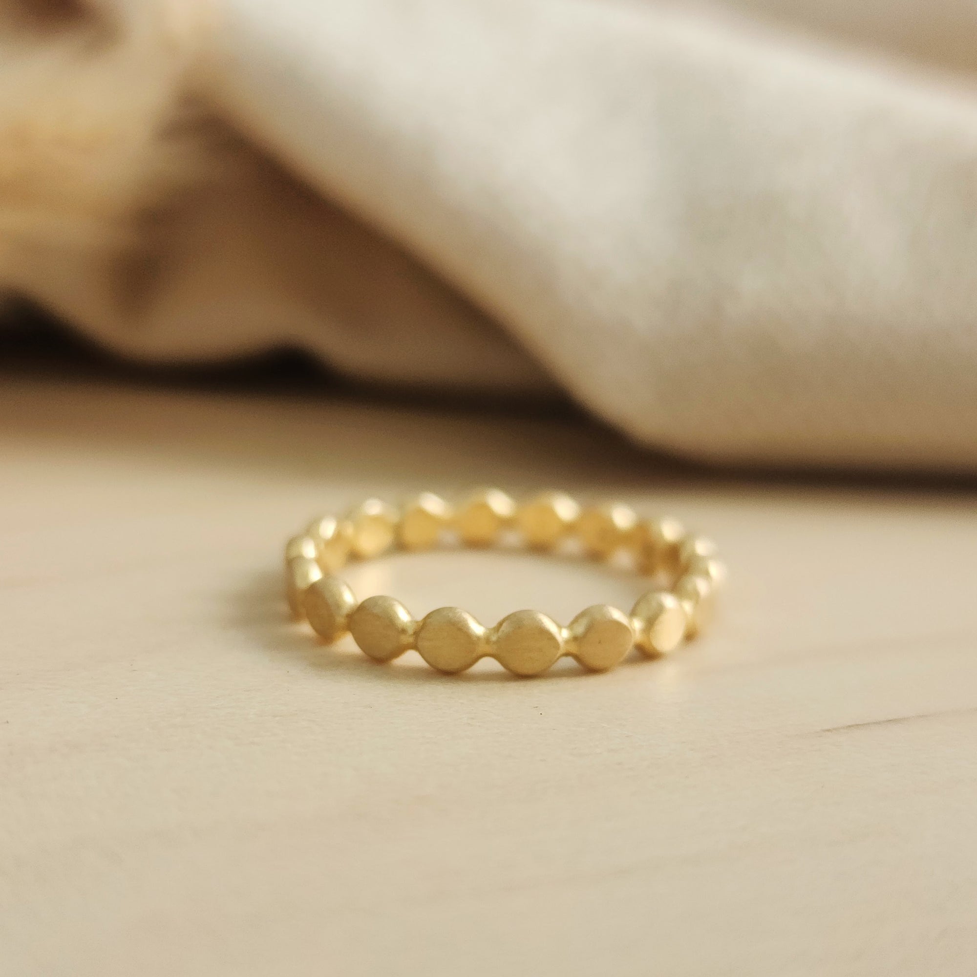 A gold-colored flat beaded stacking ring placed on a wooden surface with a neutral-colored fabric in the background.