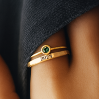 Gold ring with a green gemstone and '2024' on a person's finger against a dark fabric background
