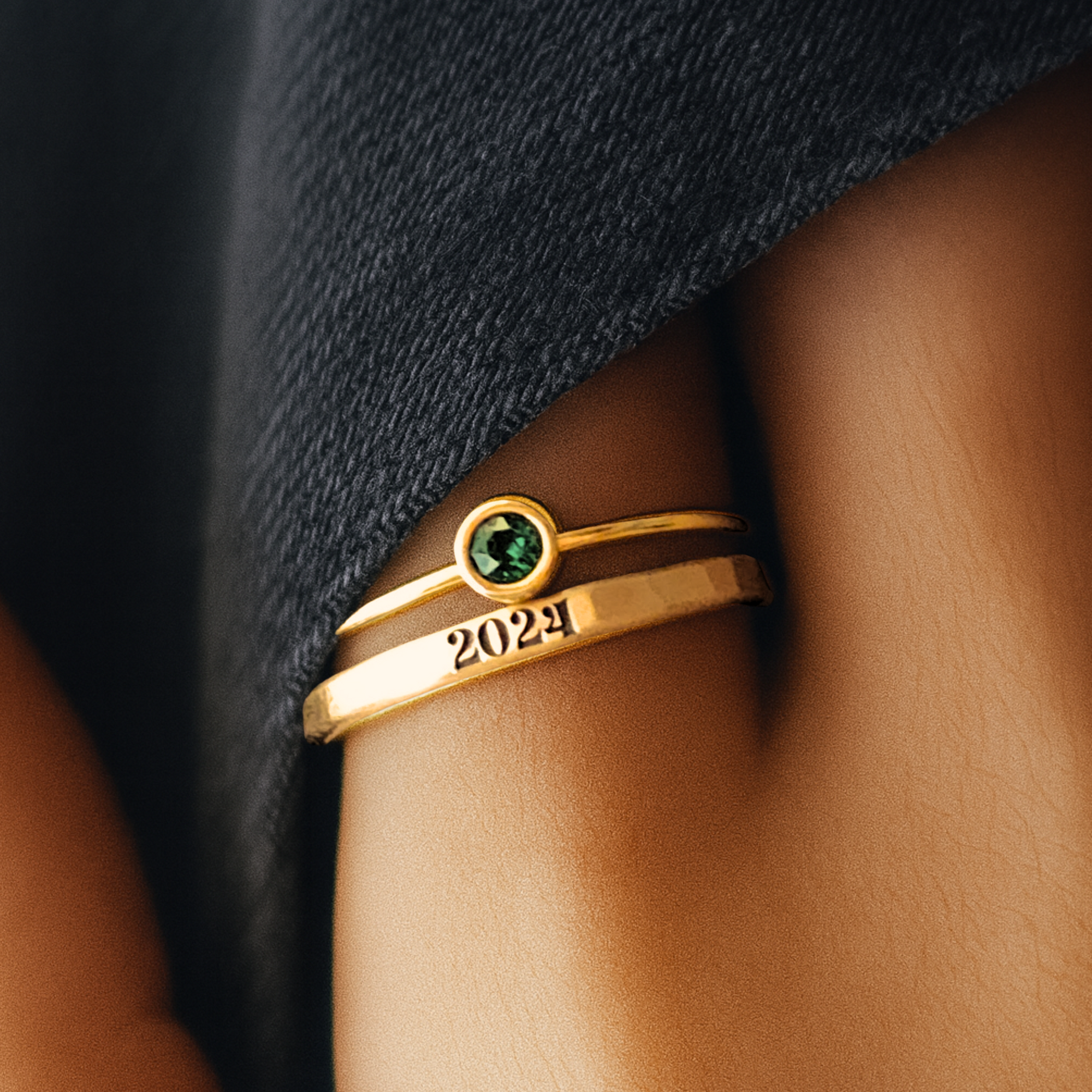 Gold ring with a green gemstone and '2024' on a person's finger against a dark fabric background
