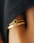 Gold ring with a green gemstone and '2024' on a person's finger against a dark fabric background
