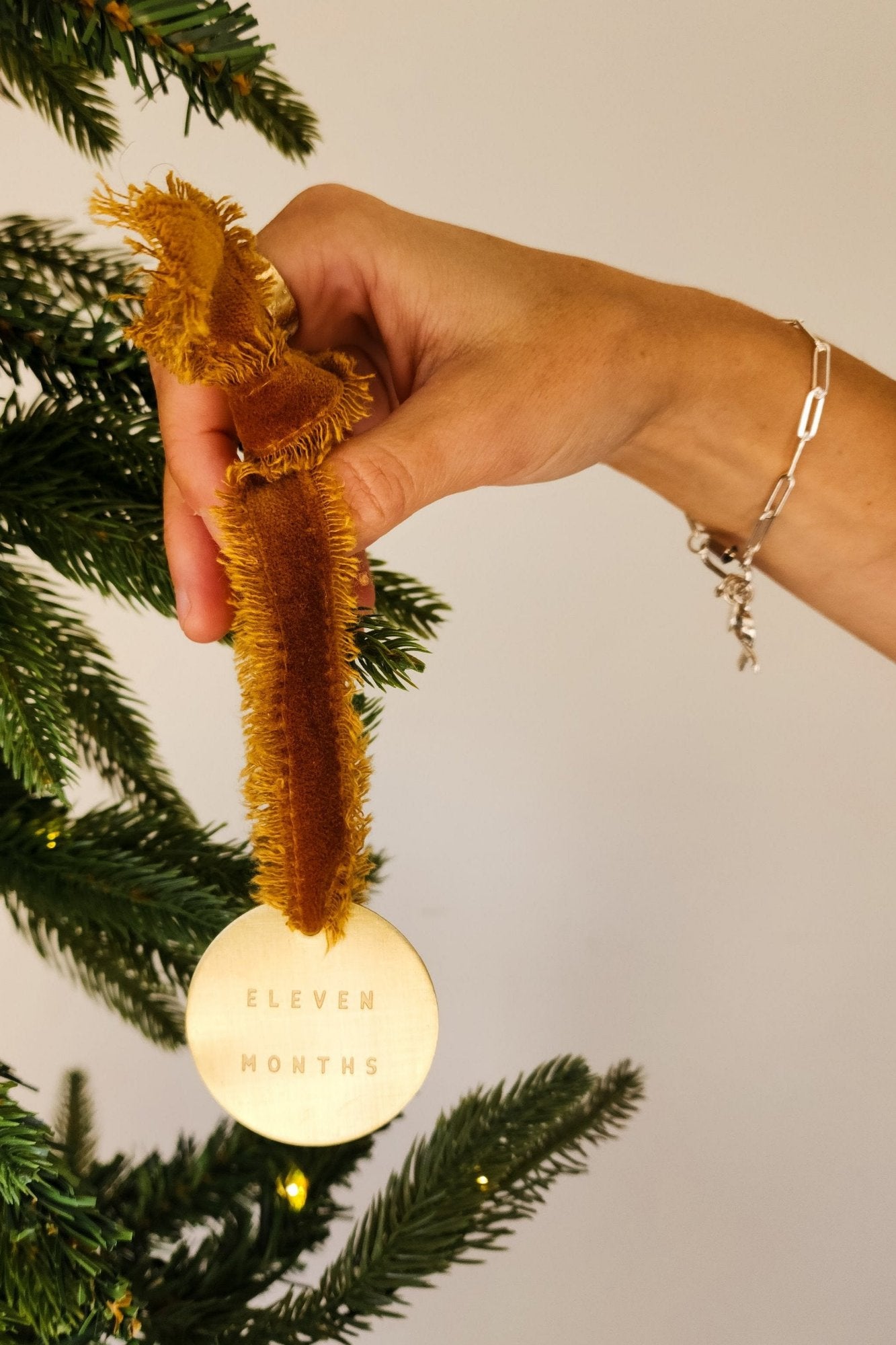 A hand holding a brass ornament with a yellow ribbon and a personalized engraving, attached to a Christmas tree.