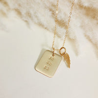Close-up of gold rectangle date necklace with angel wing charm on soft cream fabric background