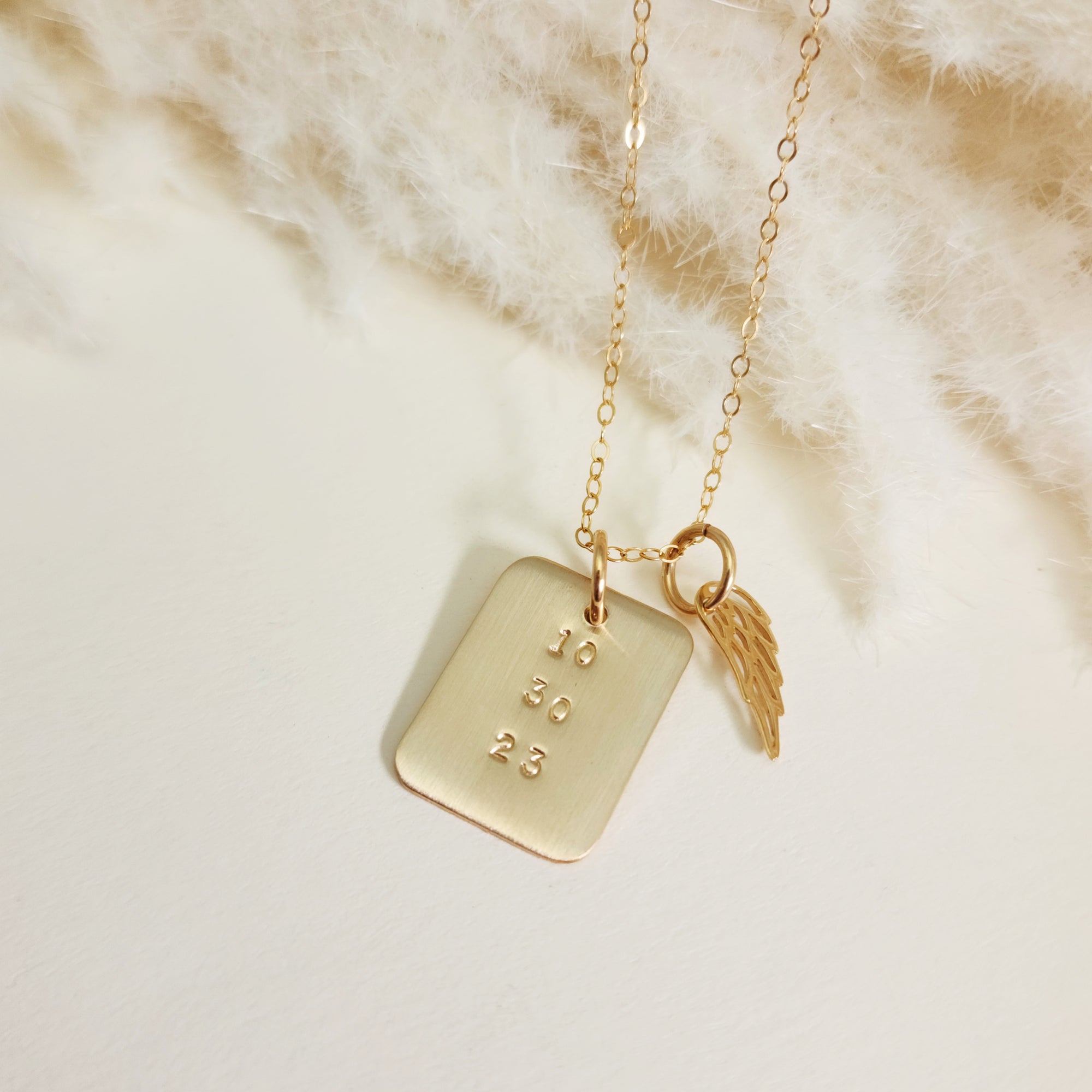 Close-up of gold rectangle date necklace with angel wing charm on soft cream fabric background