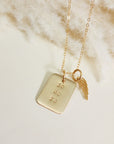 Close-up of gold rectangle date necklace with angel wing charm on soft cream fabric background