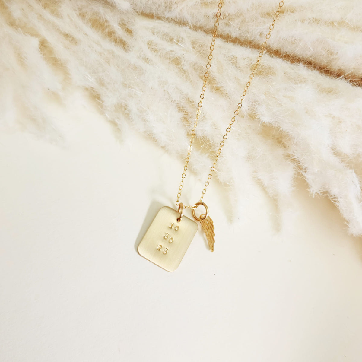 Minimal gold date necklace and angel wing charm displayed on cream textured surface