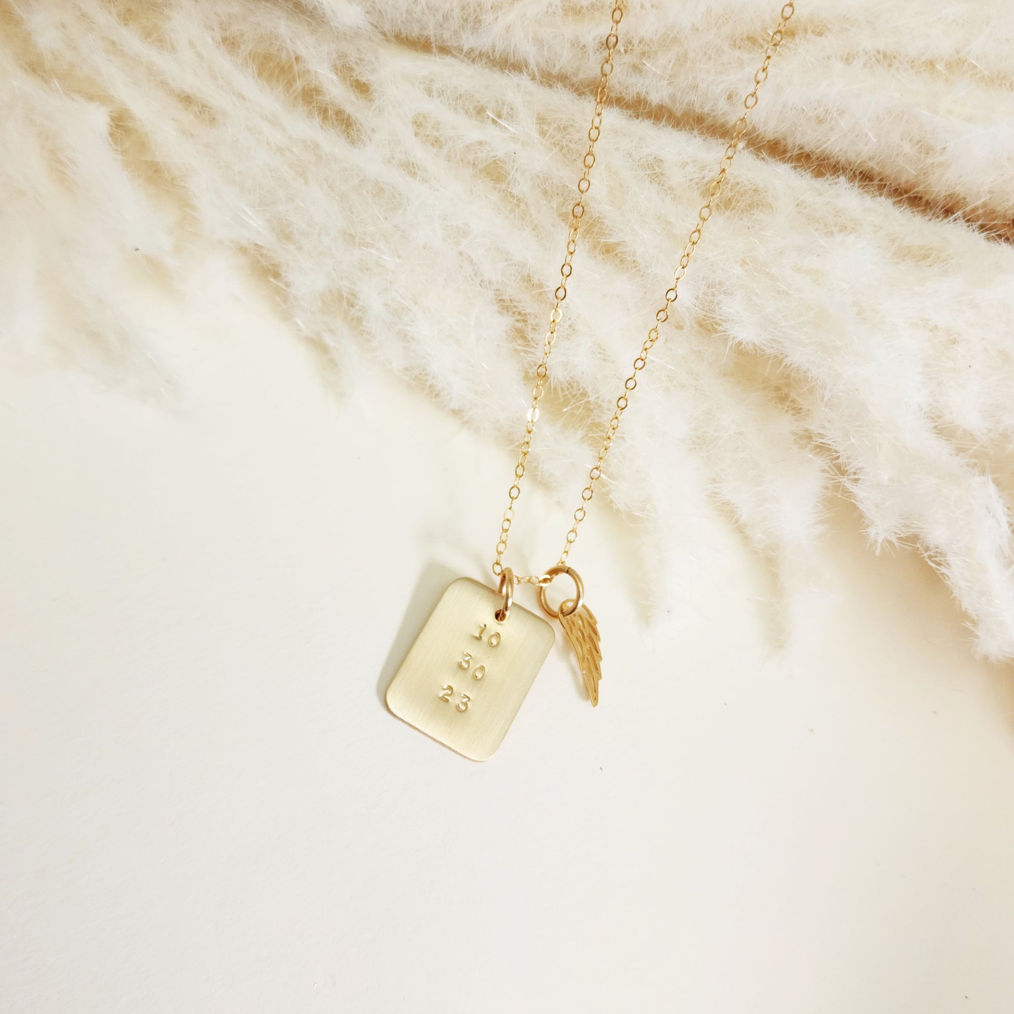Minimal gold date necklace and angel wing charm displayed on cream textured surface