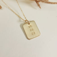 Gold date-stamped pendant necklace on gold chain displayed on a white background with gold leaf accent.