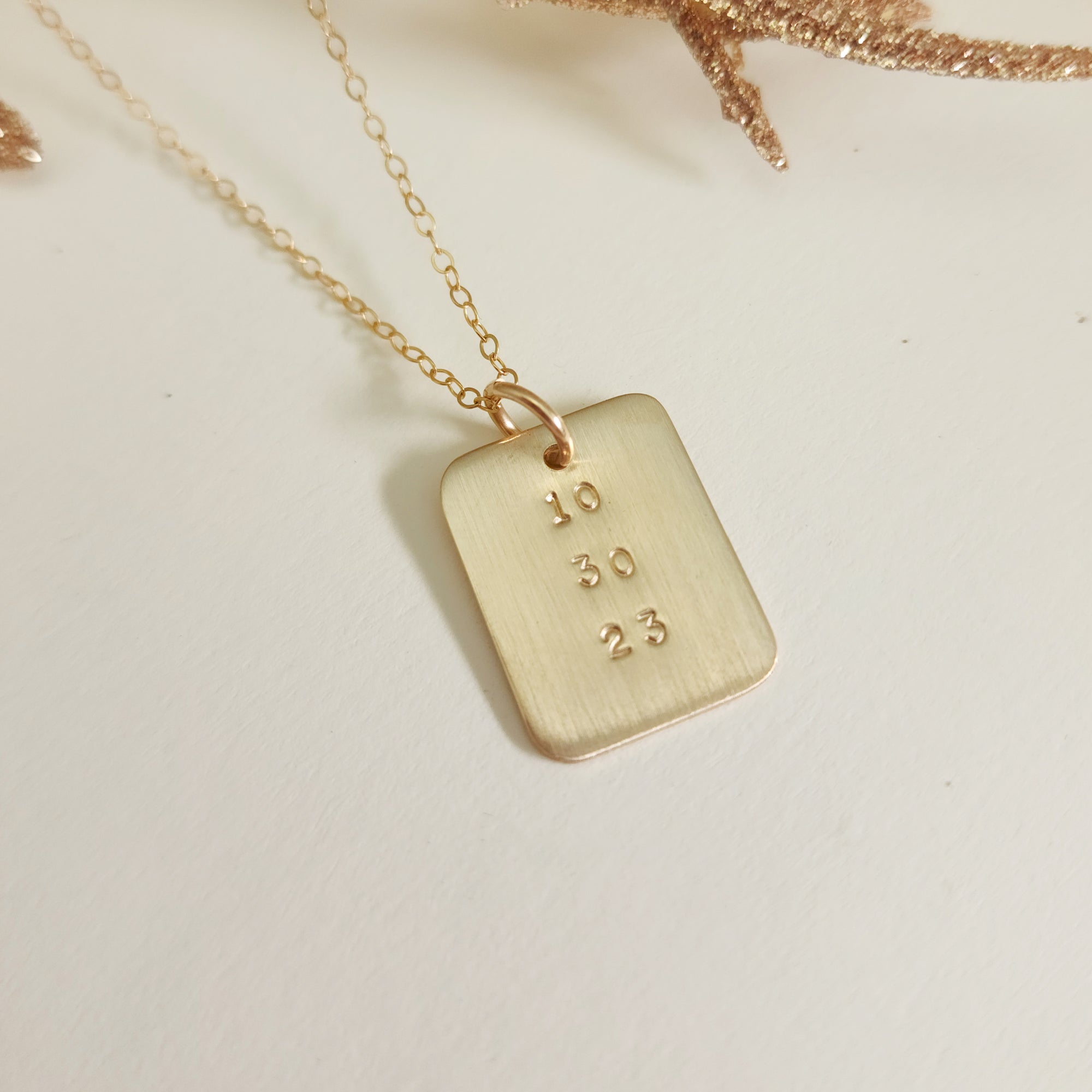 Gold date-stamped pendant necklace on gold chain displayed on a white background with gold leaf accent.