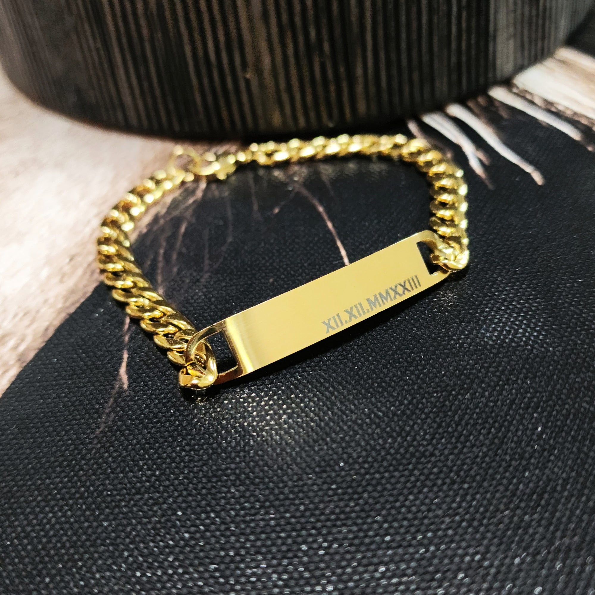 Gold chain bracelet with engraved tag on a textured black surface