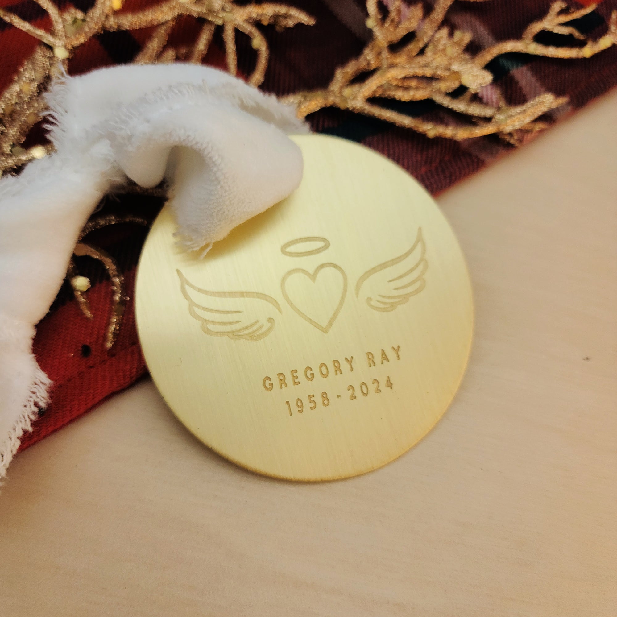 Gold engraved memorial ornament featuring angel wings, halo, and heart design on red and gold holiday fabric.