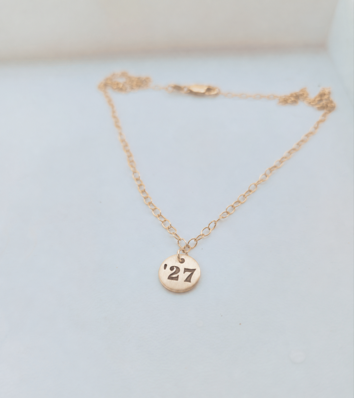 gold round pendant with '27 engraved on a cable chain laying on a surface