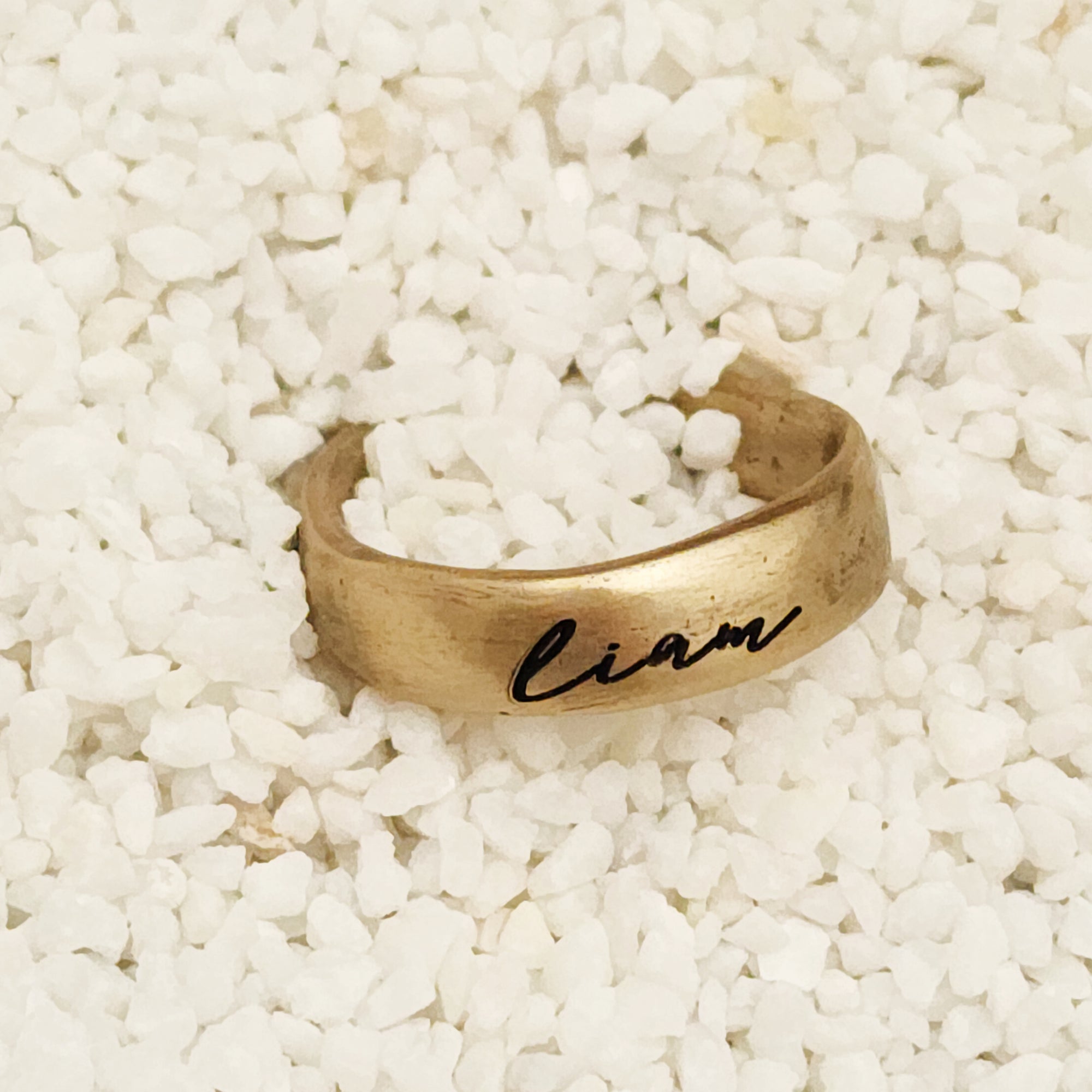 Gold ring with 'Liam' engraving on a white pebble background
