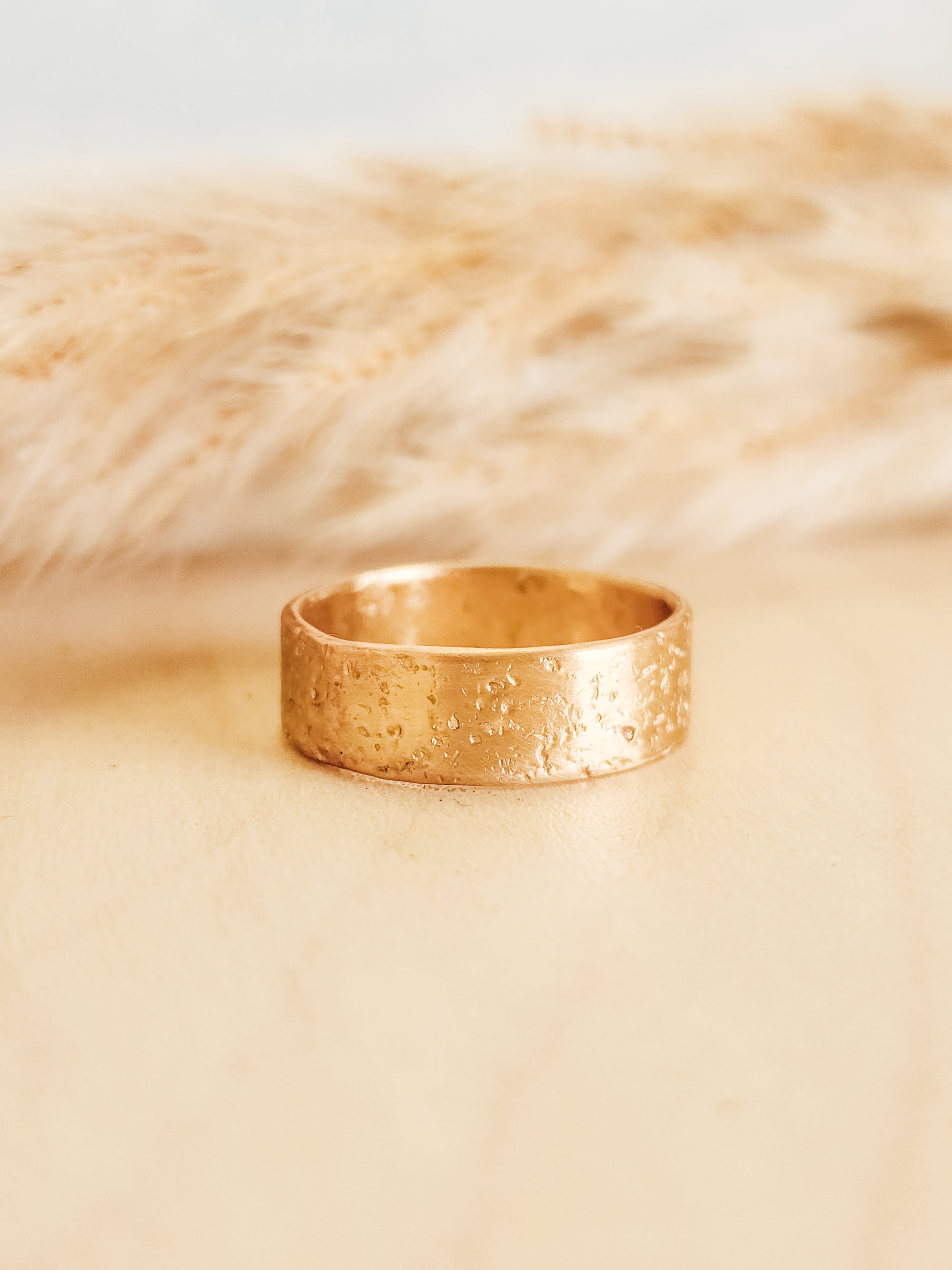 A solid gold textured ring with a thick band, placed on a beige surface next to  a decorative item.