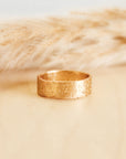 A solid gold textured ring with a thick band, placed on a beige surface next to  a decorative item.