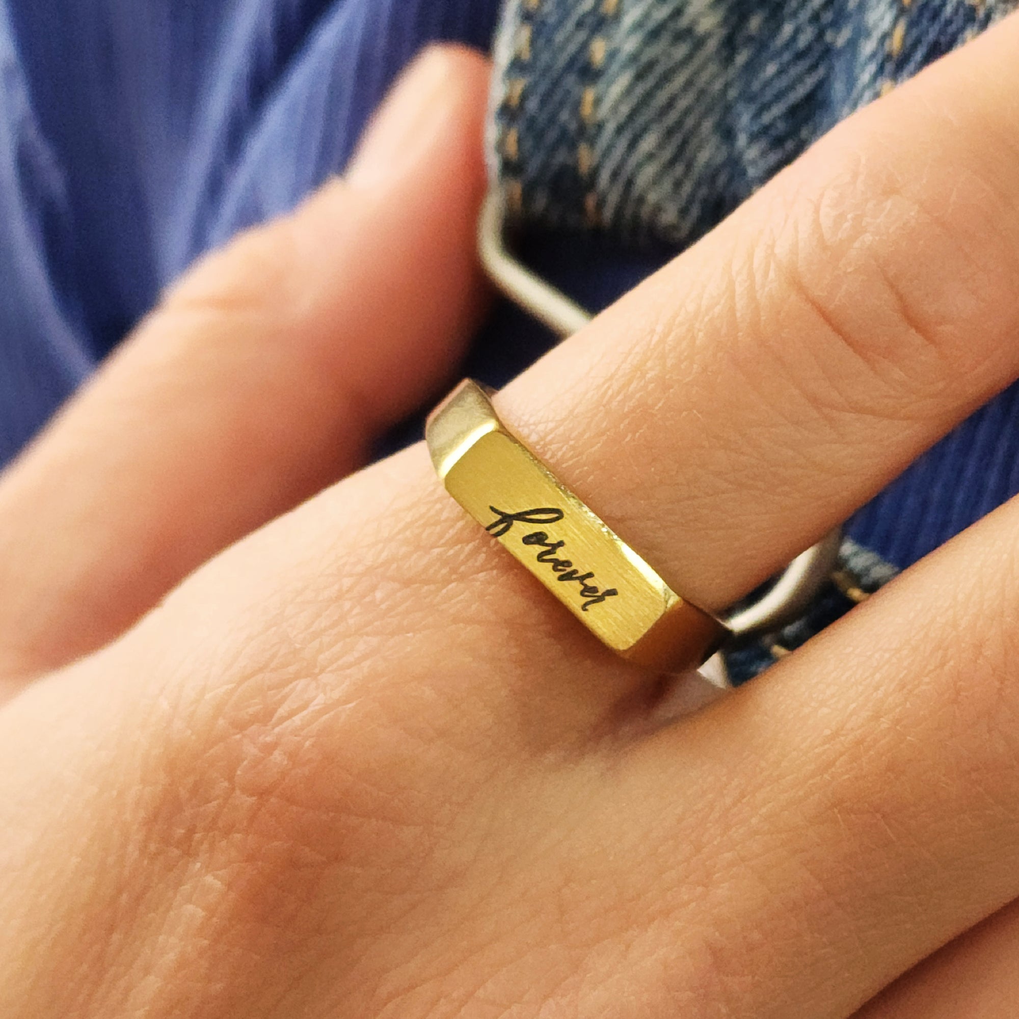 A gold-colored signet ring with custom handwriting engraved on the top surface, worn on a person's finger.