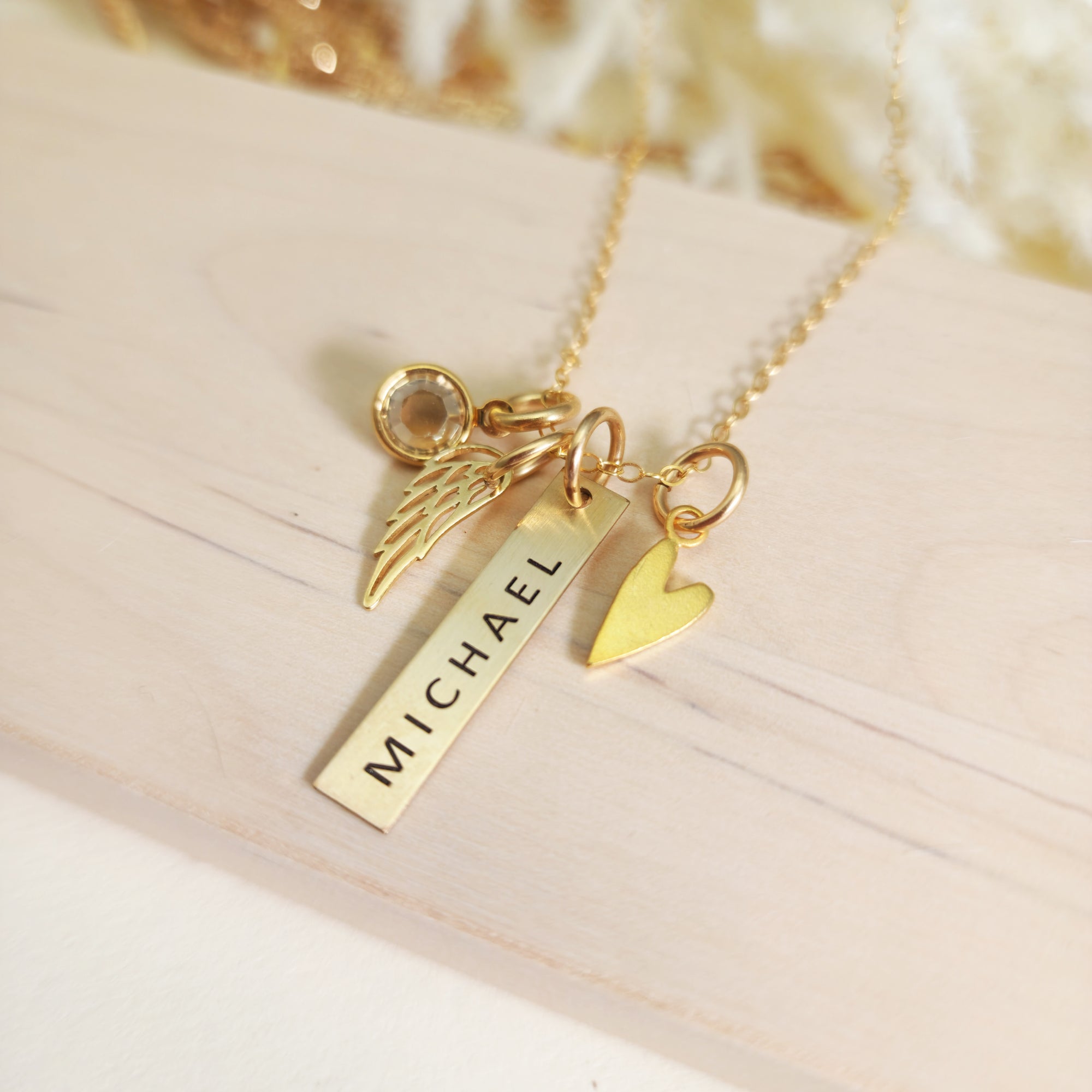 Gold memorial necklace with engraved vertical bar reading “Michael,” gold heart charm, angel wing charm, and birthstone, displayed on wood.
