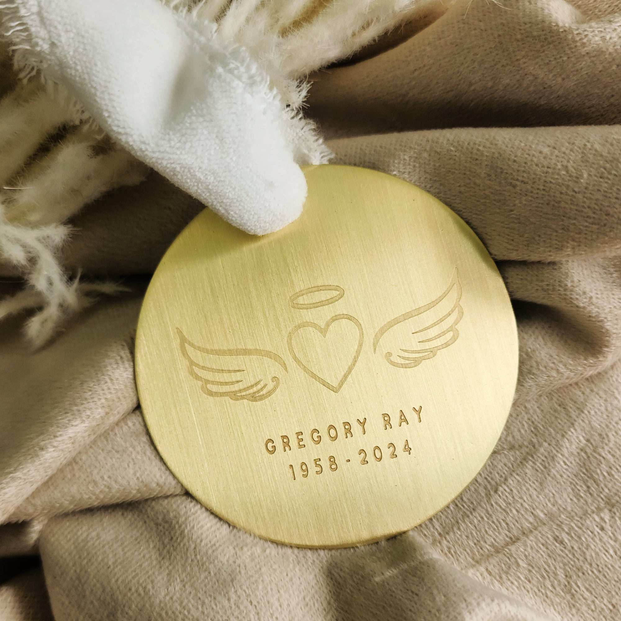 Gold memorial ornament engraved with angel wings, heart, halo, and personalized name and dates on beige fabric.
File Name: gold-memorial-or