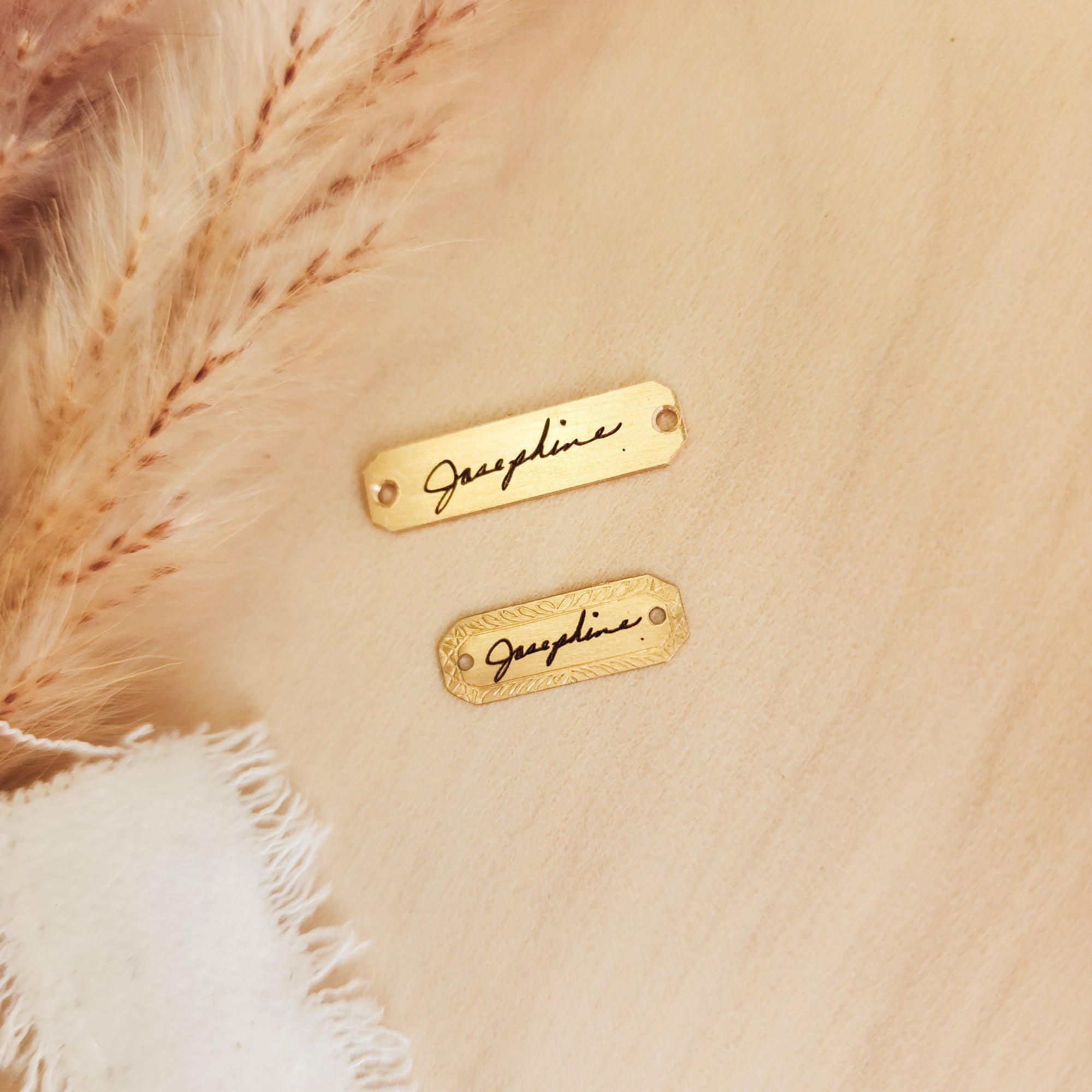 Two gold nameplates with 'Jacqueline' engraved on them, placed on a beige fabric background.