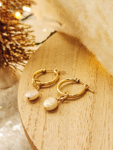 A pair of gold-colored hoop earrings with white freshwater pearl accents placed on a wooden surface with a blurred holiday-themed backdrop.