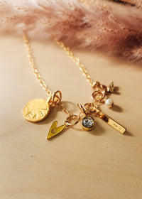 A gold-filled charm necklace with multiple charms including a heart, round tag, bar, and star, placed on a wooden surface next to a fluffy pink object.