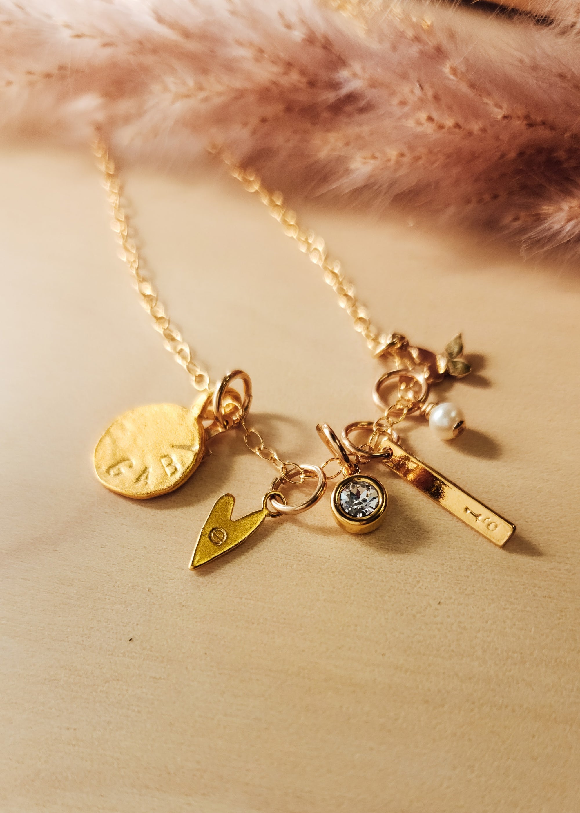A gold-filled charm necklace with multiple charms including a heart, round tag, bar, and star, placed on a wooden surface next to a fluffy pink object.