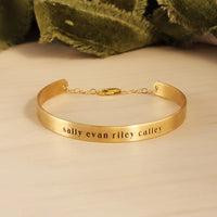 A gold-colored cuff bracelet with custom names engraved on it, displayed on a white surface with a green textured object to the side.