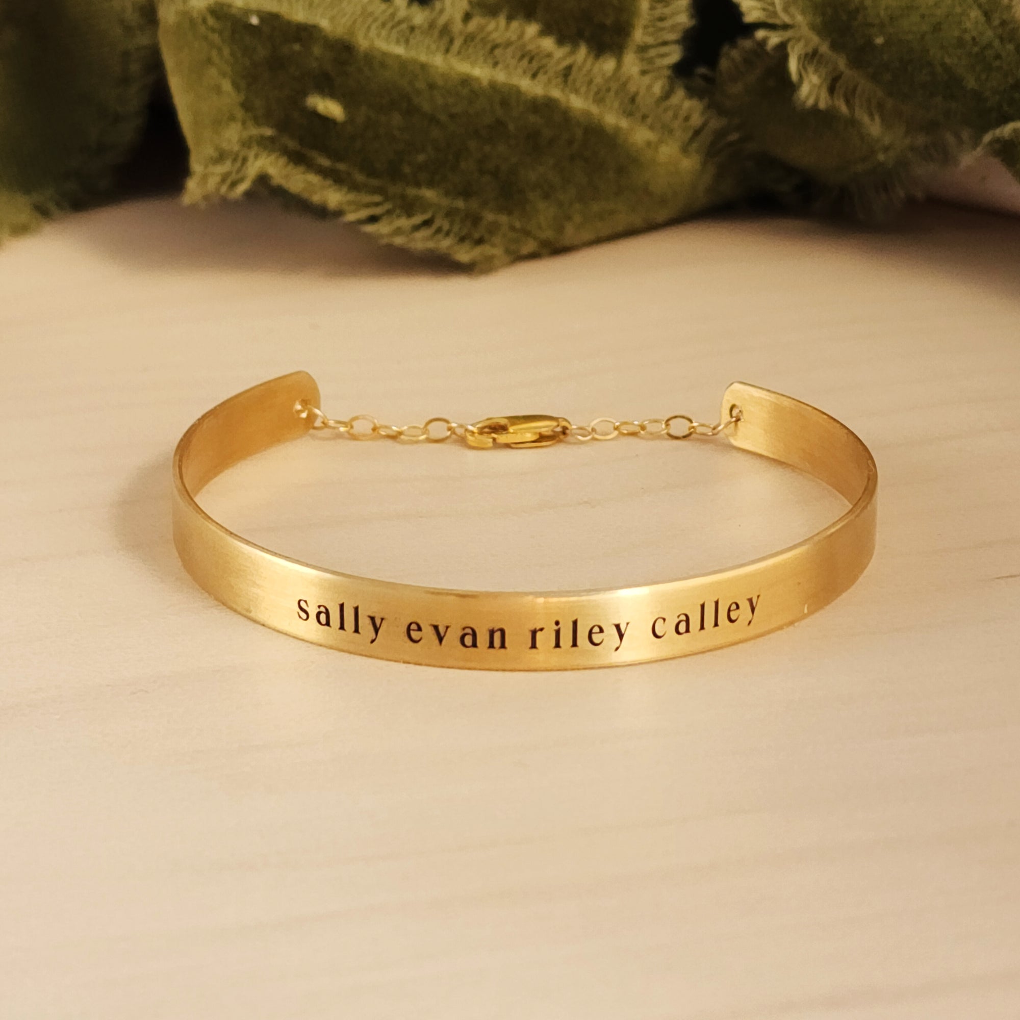 A gold-colored cuff bracelet with custom names engraved on it, displayed on a white surface with a green textured object to the side.
