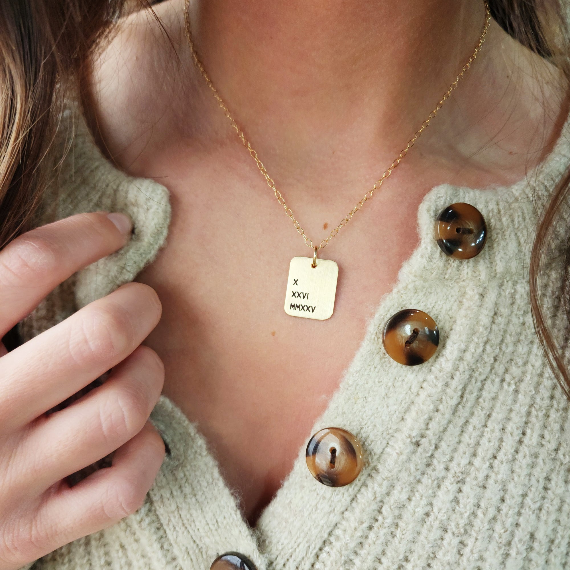 A person wearing a cardigan is showcasing a gold necklace with a rectangle pendant engraved with Roman numerals.