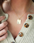 A person wearing a cardigan is showcasing a gold necklace with a rectangle pendant engraved with Roman numerals.