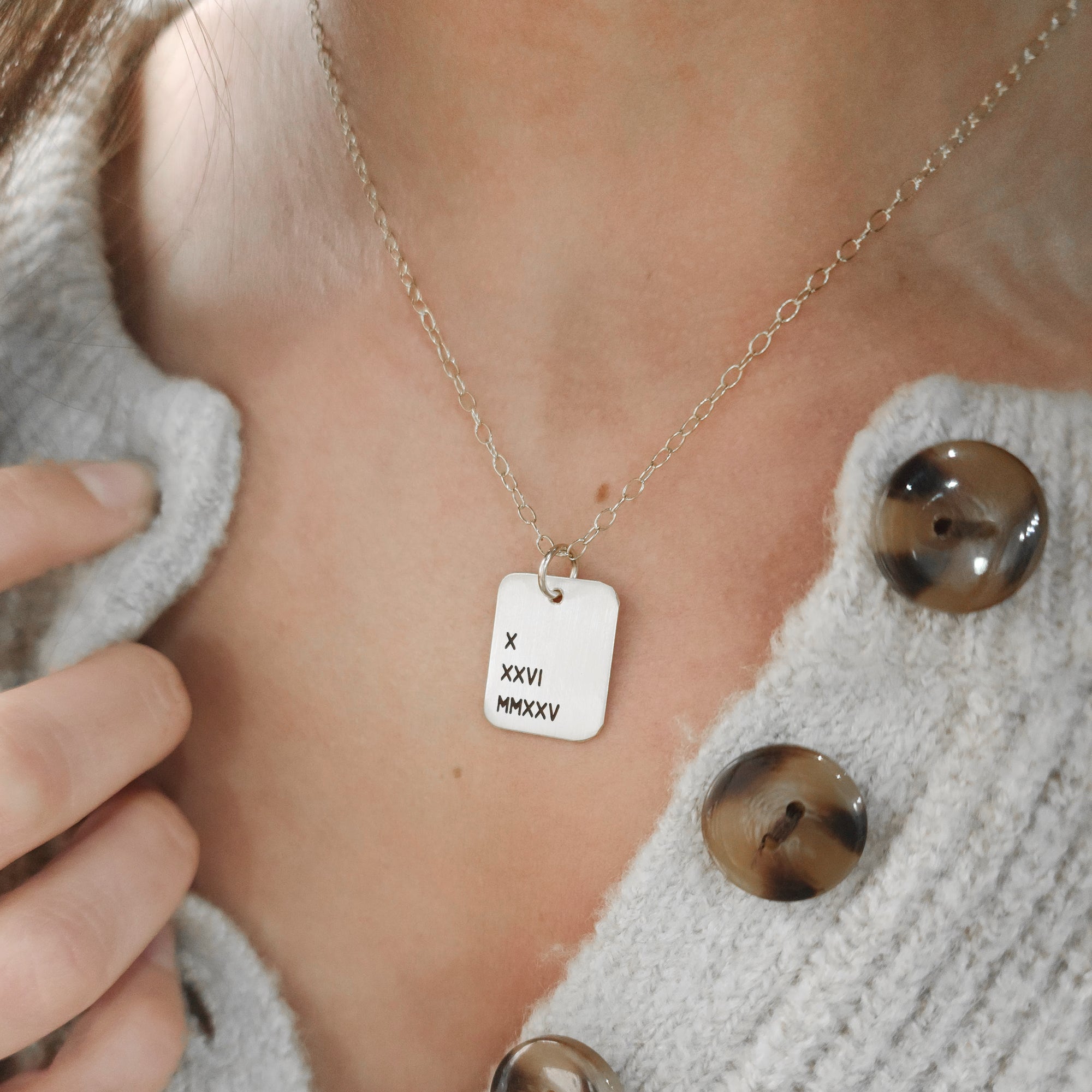 A person wearing a cardigan is showcasing a gold necklace with a rectangle pendant engraved with Roman numerals.