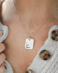 A person wearing a cardigan is showcasing a gold necklace with a rectangle pendant engraved with Roman numerals.