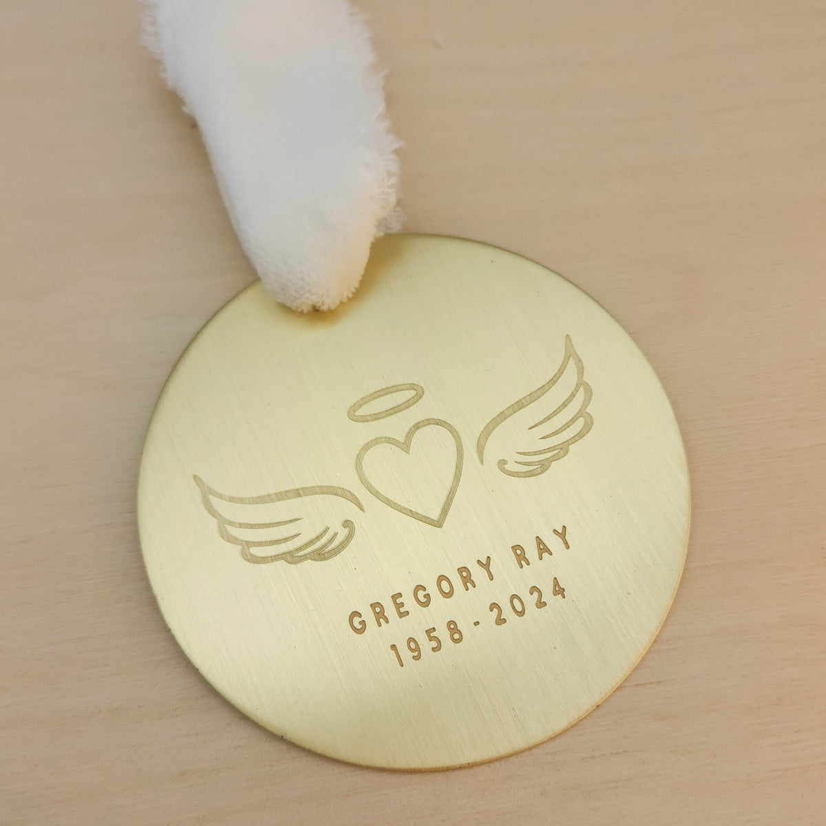 Personalized gold remembrance ornament with engraved angel wings, heart, and halo design on a light wood surface.