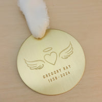 Personalized gold remembrance ornament with engraved angel wings, heart, and halo design on a light wood surface.