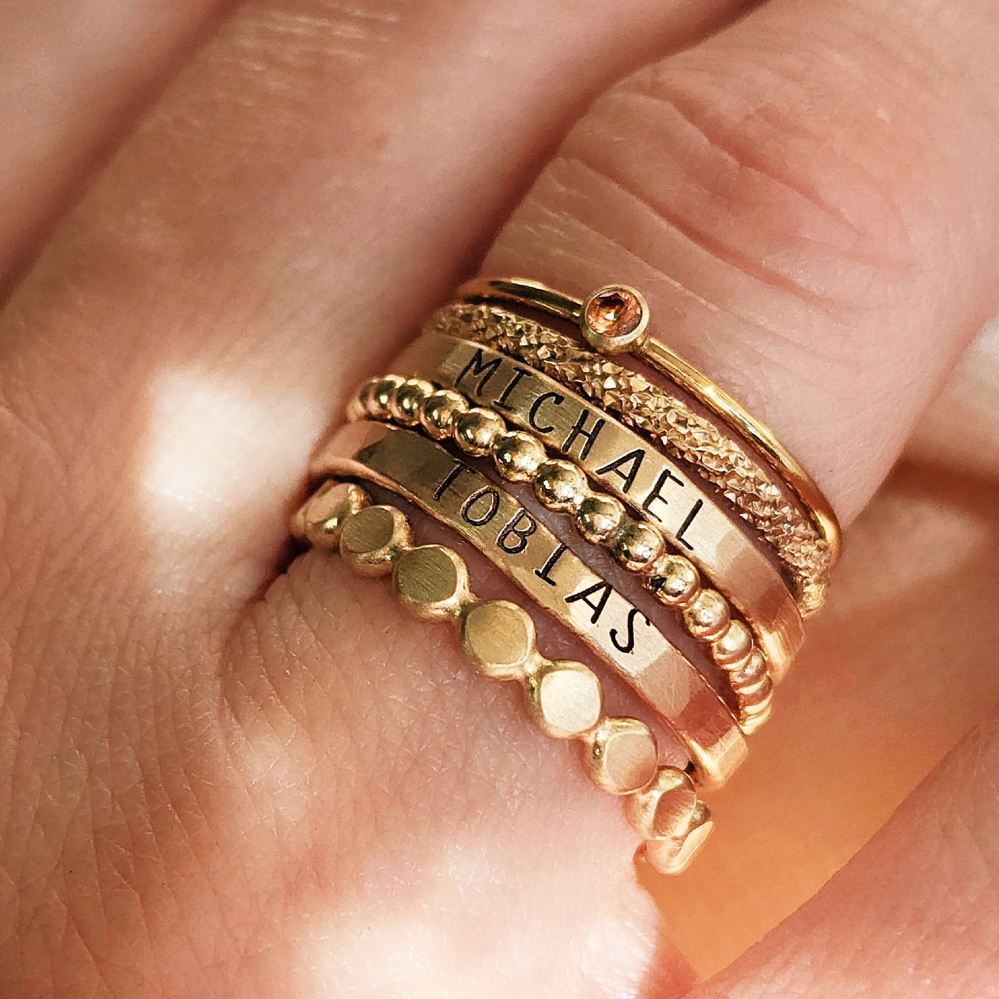 A collection of gold-colored stacking rings with varying designs, some with beaded details and personalized engravings, displayed on a person's finger.