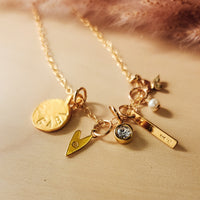 Close-up of a gold charm necklace displayed on a light wooden surface with a soft pink feather accent in the background. The necklace features several unique gold charms including: a round organic disc stamped with "GAB", a small heart with an engraved letter, a bezel-set clear gemstone, a vertical bar engraved with "10", a small pearl drop, and a tiny leaf charm. The chain is delicate and dainty, giving the piece a personalized and feminine feel.